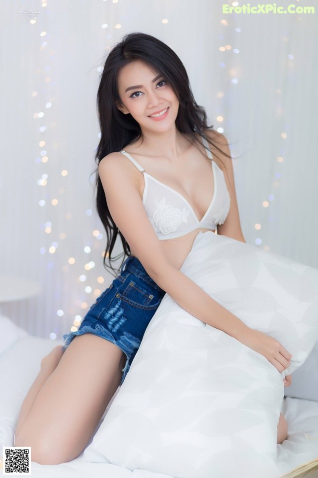 A woman sitting on top of a bed in a white bra and denim shorts.